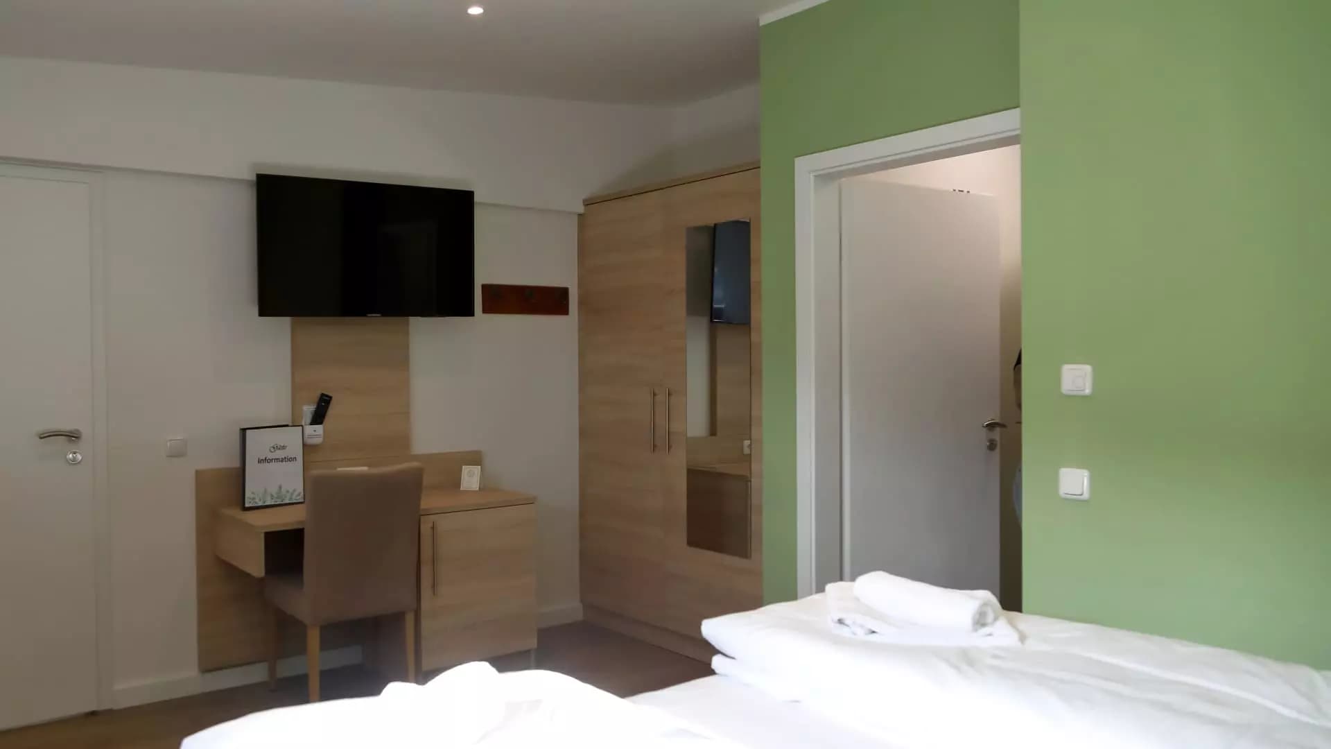 Zimmer mit Wand-TV und Schreibtisch, Blick in das grün gestaltete Badezimmer