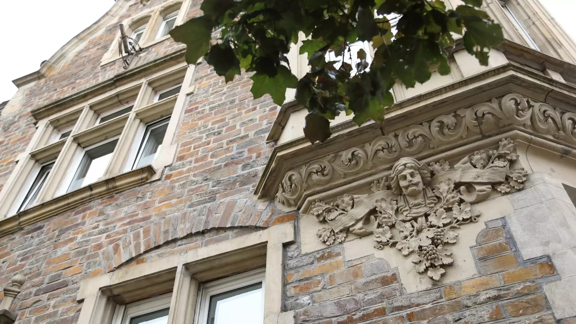 Sandstein-Ornament mit Gesichtsmaske und Weinranken an der Fassade, Blick zwischen Baumzweigen hindurch